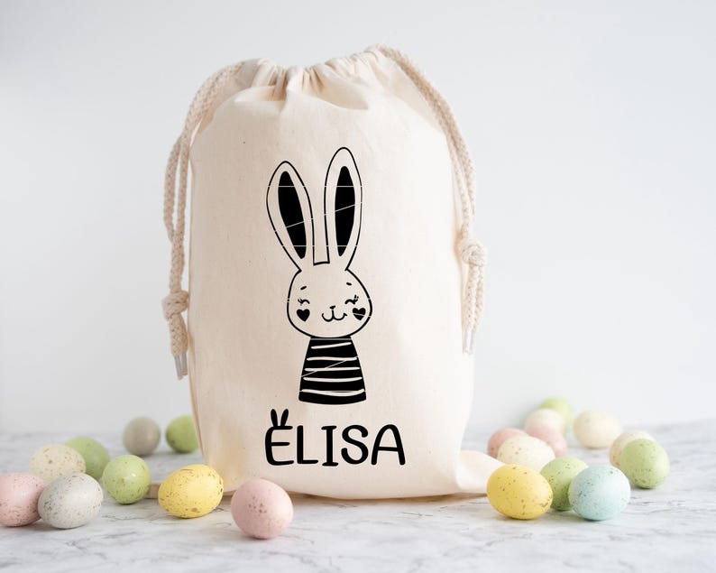 May include: A white canvas drawstring bag with a black and white bunny illustration and the name "ELISA" printed on the front. The bag is filled with pastel coloured Easter eggs.