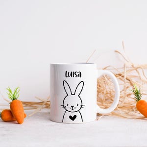 May include: White ceramic mug with a black bunny illustration and the name "Luisa" printed on it. The mug is surrounded by dried grass and two orange carrots.