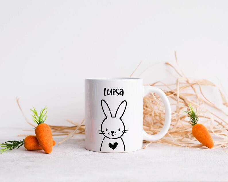 May include: White ceramic mug with a black bunny illustration and the name "Luisa" printed on it. The mug is surrounded by dried grass and two orange carrots.