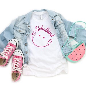 K&ouml;nnte beinhalten: Ein wei&szlig;es T-Shirt mit einem pinken Smiley und dem Text "Schulkind 20 25". Das Shirt wird mit einer hellblauen Jeansjacke und pinken Sneakern kombiniert. Eine pinkfarbene Wassermelonen-Tasche h&auml;ngt an der Jacke.