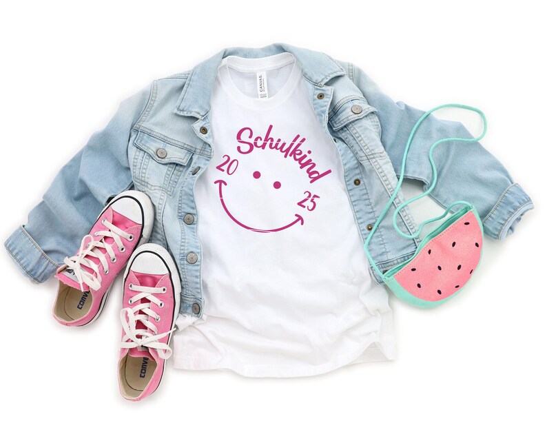 K&ouml;nnte beinhalten: Ein wei&szlig;es T-Shirt mit einem pinken Smiley und dem Text "Schulkind 20 25". Das Shirt wird mit einer hellblauen Jeansjacke und pinken Sneakern kombiniert. Eine pinkfarbene Wassermelonen-Tasche h&auml;ngt an der Jacke.