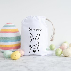 May include: White Easter gift bag with a black bunny and heart design. The bag has the name "Simone" printed on it. The bag is surrounded by pastel colored Easter eggs.