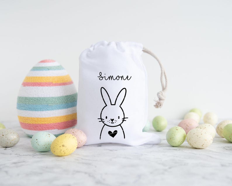 May include: White Easter gift bag with a black bunny and heart design. The bag has the name "Simone" printed on it. The bag is surrounded by pastel colored Easter eggs.