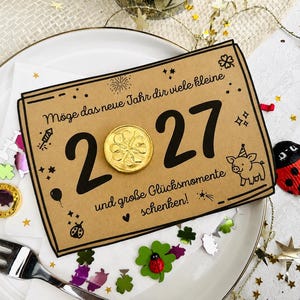 May include: A rectangular kraft paper card featuring the black text 'M&ouml;ge das neue Jahr dir viele kleine 2027 und gro&szlig;e Gl&uuml;cksmomente schenken!' with a gold foil coin replacing the zero, a small pig illustration, and festive confetti.