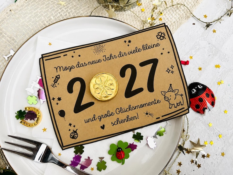 May include: A rectangular kraft paper card featuring the black text 'M&ouml;ge das neue Jahr dir viele kleine 2027 und gro&szlig;e Gl&uuml;cksmomente schenken!' with a gold foil coin replacing the zero, a small pig illustration, and festive confetti.