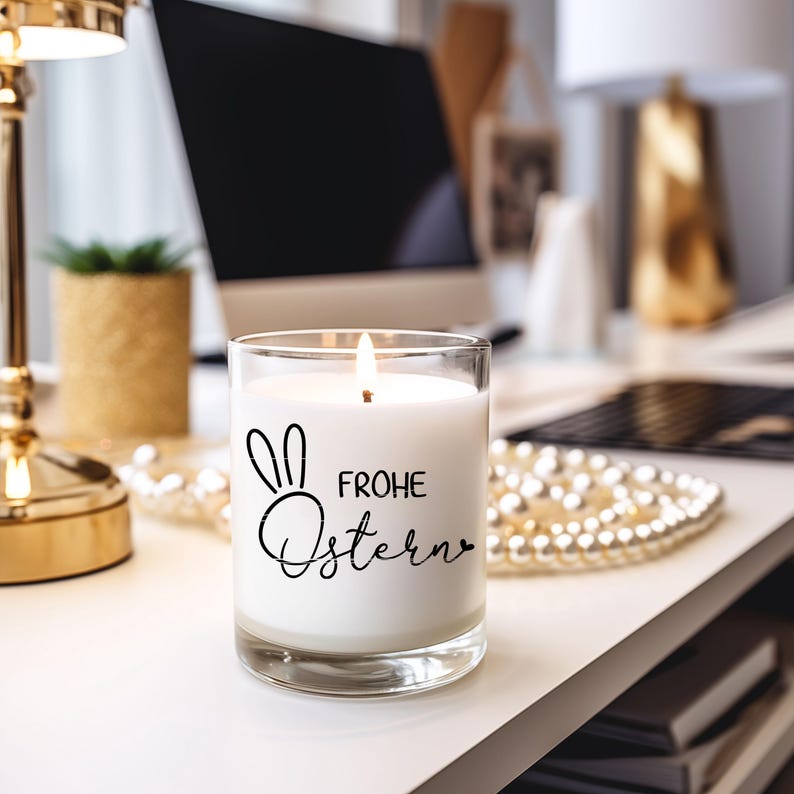Puede incluir: Una vela blanca en un tarro de vidrio transparente con un conejo negro y el texto "Frohe Ostern" en la parte delantera. La vela est&aacute; encendida y la llama es visible.