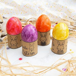 May include: Four Easter eggs decorated with different colors and patterns are displayed in brown paper holders with bunny illustrations. The holders have the text "Frohe Ostern" printed on them.