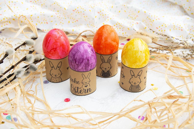 May include: Four Easter eggs decorated with different colors and patterns are displayed in brown paper holders with bunny illustrations. The holders have the text "Frohe Ostern" printed on them.