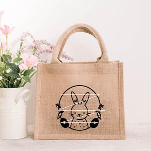 May include: A brown burlap tote bag with a black and white illustration of a bunny rabbit holding carrots. The bunny is surrounded by a circle of white string.