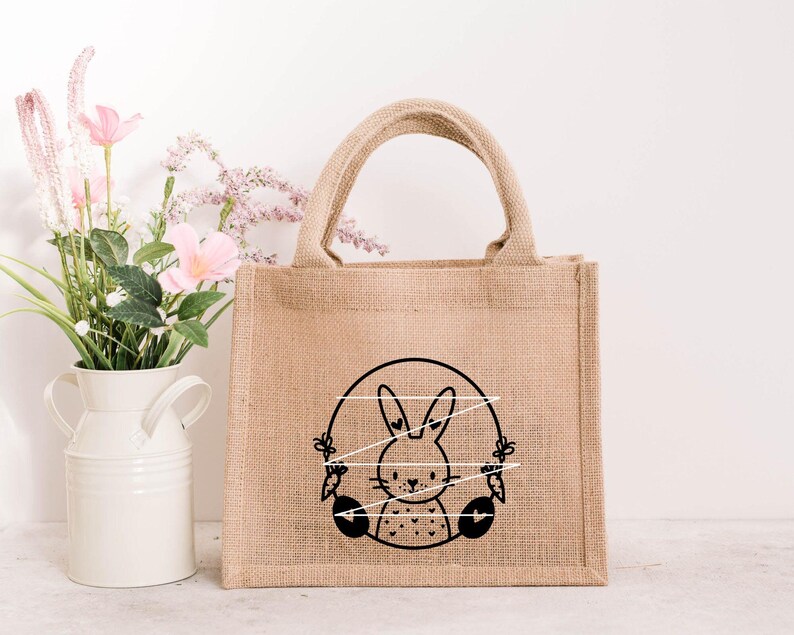 May include: A brown burlap tote bag with a black and white illustration of a bunny rabbit holding carrots. The bunny is surrounded by a circle of white string.