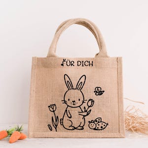 May include: A beige burlap tote bag featuring a black illustration of a rabbit holding a flower, a bee, and a ladybug. The text "F&Uuml;R DICH" is printed above the rabbit. The bag has two handles and is on a white surface.