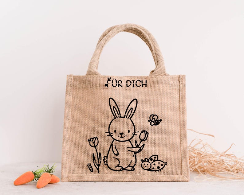 May include: A beige burlap tote bag featuring a black illustration of a rabbit holding a flower, a bee, and a ladybug. The text "F&Uuml;R DICH" is printed above the rabbit. The bag has two handles and is on a white surface.
