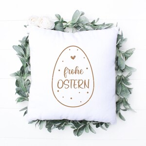 K&ouml;nnte beinhalten: Ein wei&szlig;es Kissen mit einem braunen Osterei-Design und dem Text "frohe Ostern".