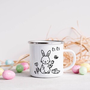 May include: White enamel mug featuring a black illustration of a rabbit holding a flower, with a butterfly and ladybug. The mug has a silver rim and a white handle. Colourful Easter eggs are scattered around the mug.
