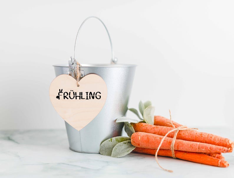 May include: A silver metal bucket with a wooden heart-shaped tag that reads "FR&Uuml;HLING" in black. A bundle of orange carrots tied with twine and green leaves rests beside the bucket, suggesting a spring or Easter theme.
