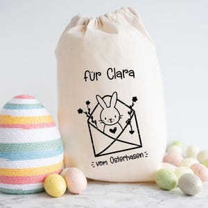 May include: A white canvas bag with black text that reads "f&uuml;r Clara = vom Osterhasen =."  The bag has a black and white illustration of a bunny in an envelope with flowers. A striped Easter egg is in the foreground.