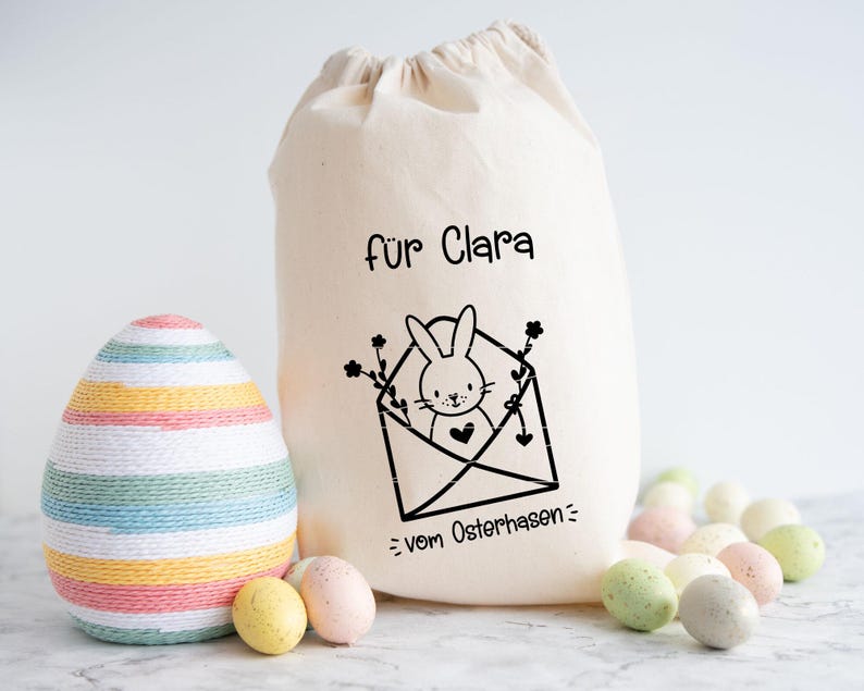 May include: A white canvas bag with black text that reads "f&uuml;r Clara = vom Osterhasen =."  The bag has a black and white illustration of a bunny in an envelope with flowers. A striped Easter egg is in the foreground.