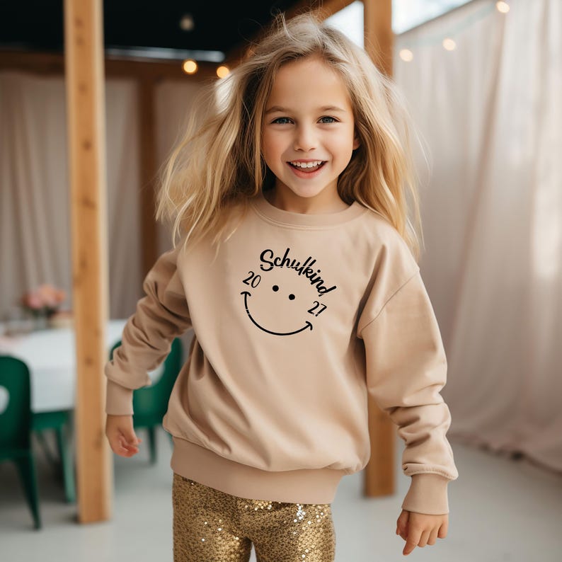 K&ouml;nnte beinhalten: Ein beiger Pullover mit einem schwarzen Smiley und dem Text "Schulkind 20 21" auf der Vorderseite. Der Pullover wird von einem jungen M&auml;dchen mit langen blonden Haaren getragen.