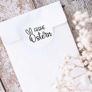 May include: White envelope with a round sticker that says "Frohe Ostern" with a bunny rabbit silhouette. The envelope is on a whitewashed wood surface with white flowers.