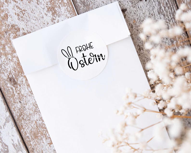 May include: White envelope with a round sticker that says "Frohe Ostern" with a bunny rabbit silhouette. The envelope is on a whitewashed wood surface with white flowers.