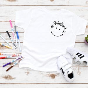 K&ouml;nnte beinhalten: Wei&szlig;es T-Shirt mit einem schwarzen Smiley und dem Text "Schulkind 20 27" auf der Vorderseite. Das Shirt liegt auf einer wei&szlig;en Holzoberfl&auml;che mit bunten Markern und einem Paar wei&szlig;en Sneakers mit schwarzen Streifen.