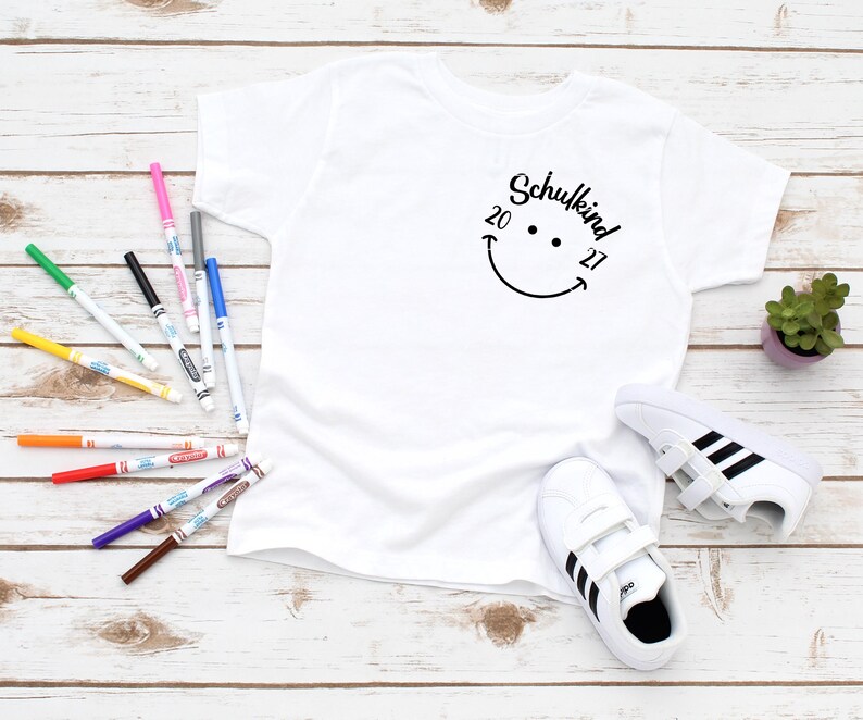 K&ouml;nnte beinhalten: Wei&szlig;es T-Shirt mit einem schwarzen Smiley und dem Text "Schulkind 20 27" auf der Vorderseite. Das Shirt liegt auf einer wei&szlig;en Holzoberfl&auml;che mit bunten Markern und einem Paar wei&szlig;en Sneakers mit schwarzen Streifen.