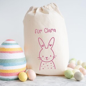 May include: A cream-colored drawstring bag with a pink bunny illustration and the text "f&uuml;r Clara." An Easter egg wrapped in colorful yarn sits next to the bag, along with several speckled Easter eggs in pastel colors.