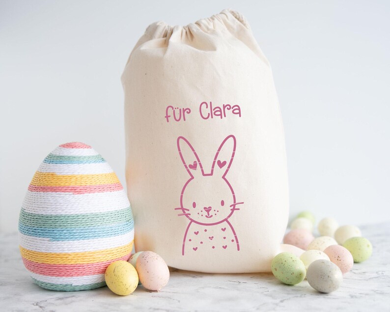 May include: A cream-colored drawstring bag with a pink bunny illustration and the text "f&uuml;r Clara." An Easter egg wrapped in colorful yarn sits next to the bag, along with several speckled Easter eggs in pastel colors.