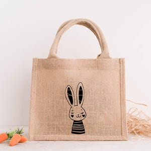 Op de afbeelding: Een beige jute draagtas met een zwart-witte konijnenillustratie. Het konijn draagt een gestreept shirt en heeft twee harten als ogen.