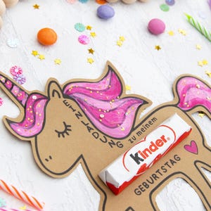 May include: A brown cardboard unicorn cutout with pink glitter mane and tail. The unicorn has a slot for a chocolate bar, which is a Kinder chocolate bar with the text "Kinder" and "Geburtstag" on it. The unicorn is on a white background with confetti.