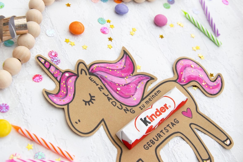 May include: A brown cardboard unicorn cutout with pink glitter mane and tail. The unicorn has a slot for a chocolate bar, which is a Kinder chocolate bar with the text "Kinder" and "Geburtstag" on it. The unicorn is on a white background with confetti.