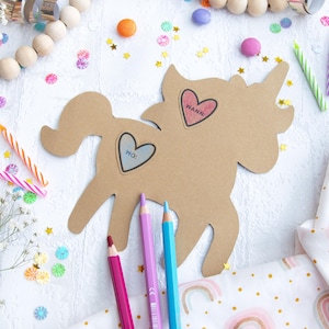 May include: A brown cardboard unicorn cutout with two heart-shaped stickers that say "Wanna" and "Pic." The unicorn is surrounded by colorful confetti, candles, and pencils.