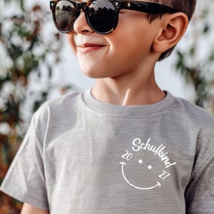 K&ouml;nnte beinhalten: Ein graues T-Shirt mit einem wei&szlig;en Smiley-Design, auf dem "Schulkind 20 27" steht. Ein Kind tr&auml;gt das Shirt und eine Sonnenbrille.