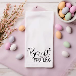 May include: White kitchen towel with the text "Bereit fur den Fr&uuml;hling" (Ready for Spring) in black cursive lettering with a small butterfly graphic. The towel is surrounded by pastel pink and white candy eggs.