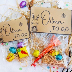 Op de afbeelding: Twee bruine papieren zakken met konijnenillustraties en de tekst "Ostern to go" erop. De zakken zijn gevuld met paaslekkernijen, waaronder chocolade-eieren, een wortel en een kleine paraplu.