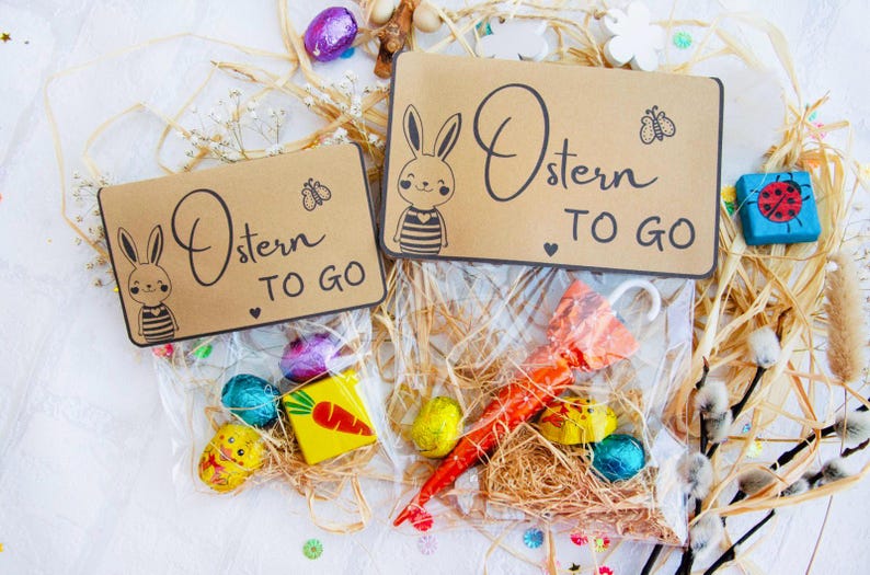 Op de afbeelding: Twee bruine papieren zakken met konijnenillustraties en de tekst "Ostern to go" erop. De zakken zijn gevuld met paaslekkernijen, waaronder chocolade-eieren, een wortel en een kleine paraplu.