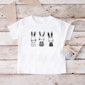 May include: White t-shirt featuring a black and white graphic of three bunnies wearing different outfits. The bunnies are wearing striped shirts, a polka dot shirt, and a plain shirt.