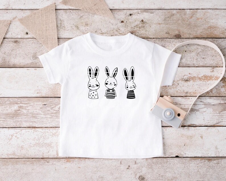 May include: White t-shirt featuring a black and white graphic of three bunnies wearing different outfits. The bunnies are wearing striped shirts, a polka dot shirt, and a plain shirt.