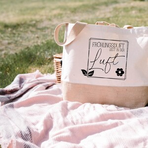 May include: A white tote bag with a brown jute bottom. The bag has black text that reads "Fr&uuml;hlingsduft liegt in der Luft" with a small flower graphic.