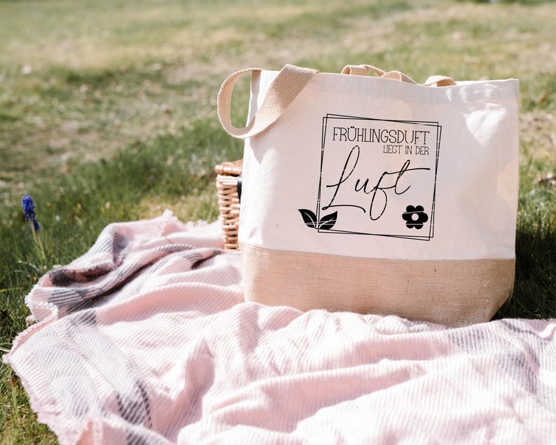 May include: A white tote bag with a brown jute bottom. The bag has black text that reads "Fr&uuml;hlingsduft liegt in der Luft" with a small flower graphic.