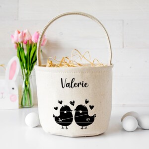 May include: A white canvas Easter basket with a handle, personalized with the name "Valerie". The basket features a black and white illustration of two birds facing each other with hearts around them.