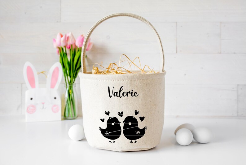 May include: A white canvas Easter basket with a handle, personalized with the name "Valerie". The basket features a black and white illustration of two birds facing each other with hearts around them.