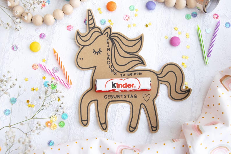 May include: A brown cardboard unicorn cutout with a Kinder chocolate bar inside. The unicorn has a black outline and the words "Einladung zu meinem Geburtstag" written on it. The unicorn is surrounded by colorful confetti and candles.