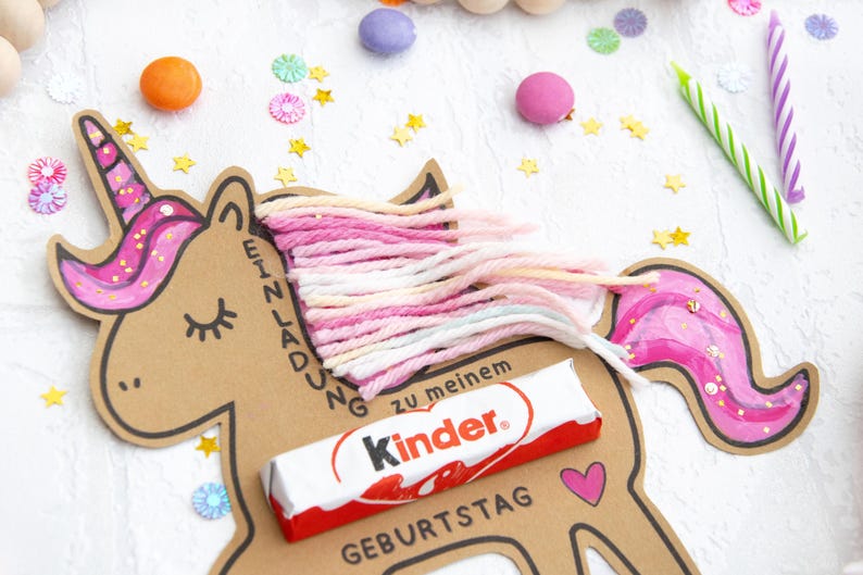 May include: A brown cardboard unicorn with pink glitter and a pink, white, and peach yarn mane. The unicorn has a Kinder chocolate bar attached to it and the text "Einladung zu meinem Geburtstag".