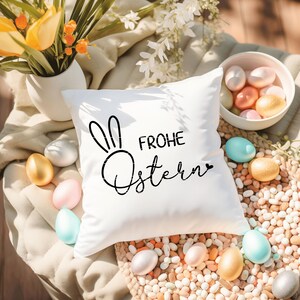 Puede incluir: Almohada blanca con texto negro que dice "Frohe Ostern" y una silueta de oreja de conejo negra. La almohada est&aacute; rodeada de huevos de Pascua de color pastel y una cesta de huevos.