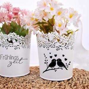 May include: Two white metal flower pots with lace designs. One pot has the text "Fr&uuml;hlingszeit" and the other has a silhouette of two birds on a branch with hearts. Both pots are filled with artificial flowers.