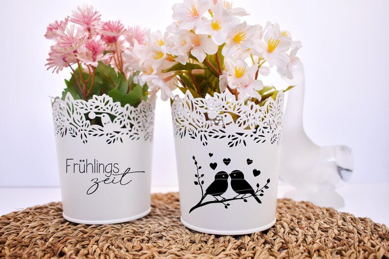May include: Two white metal flower pots with lace designs. One pot has the text "Fr&uuml;hlingszeit" and the other has a silhouette of two birds on a branch with hearts. Both pots are filled with artificial flowers.