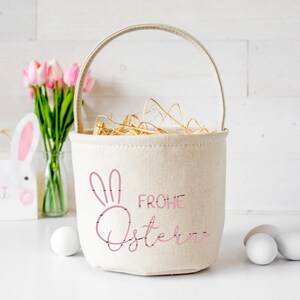 Puede incluir: Una cesta de Pascua de lona blanca con un conejito rosa y el texto "Frohe Ostern" en rosa. La cesta est&aacute; llena de paja y hay flores rosas y huevos de Pascua blancos en el fondo.