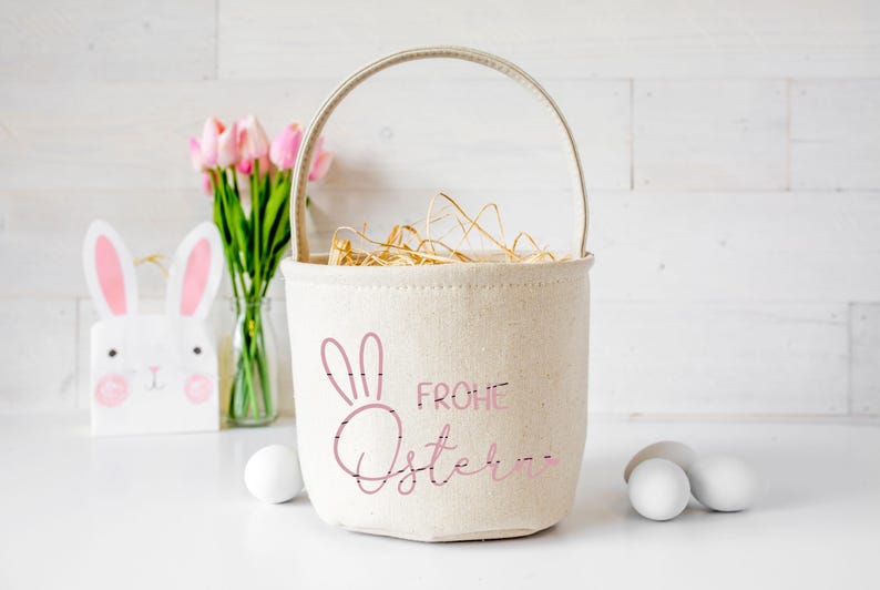 Puede incluir: Una cesta de Pascua de lona blanca con un conejito rosa y el texto "Frohe Ostern" en rosa. La cesta est&aacute; llena de paja y hay flores rosas y huevos de Pascua blancos en el fondo.