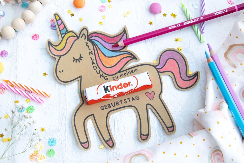 May include: A brown paper unicorn cutout with a rainbow mane and tail. The unicorn has a chocolate bar with the text "Kinder" on it, and the words "Einladung zu meinem Geburtstag" written on the side.